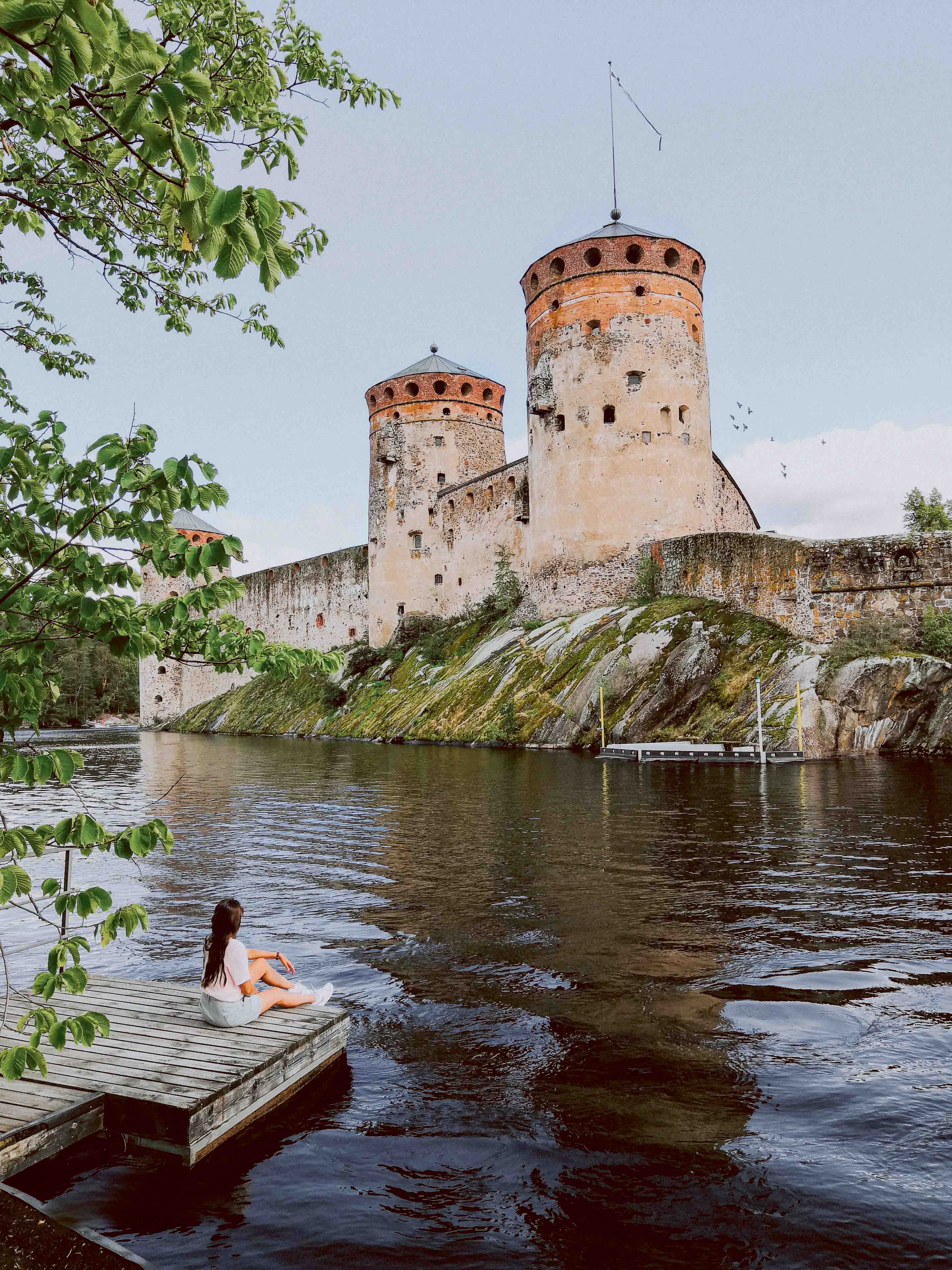 a woman sitting on a pier staring at Olavinlinna castle