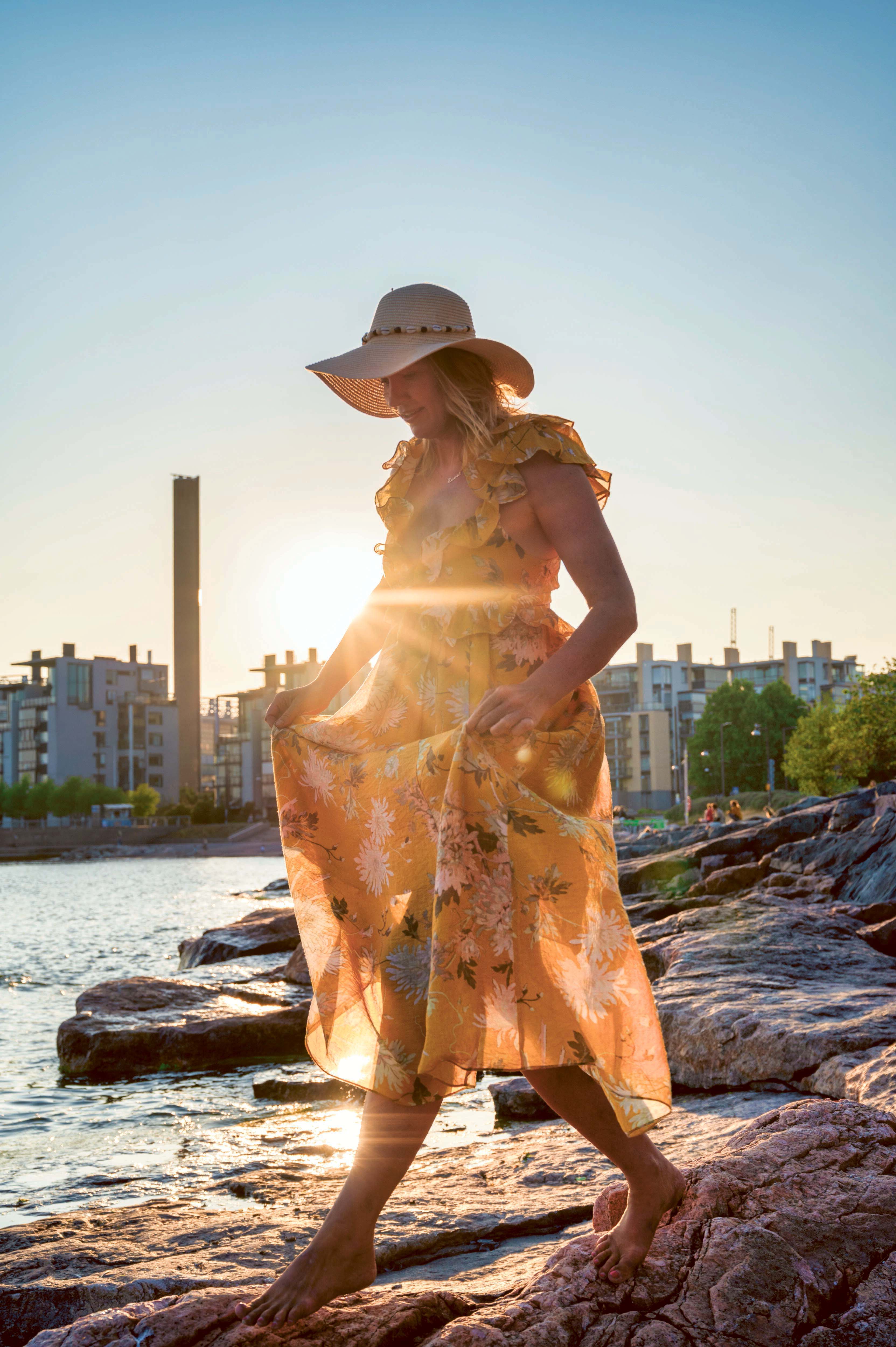 Eine Frau in einem gelben Sommerkleid steht auf einem Felsen am Ufer von Kaivopuisto in der urbanen Landschaft von Helsinki