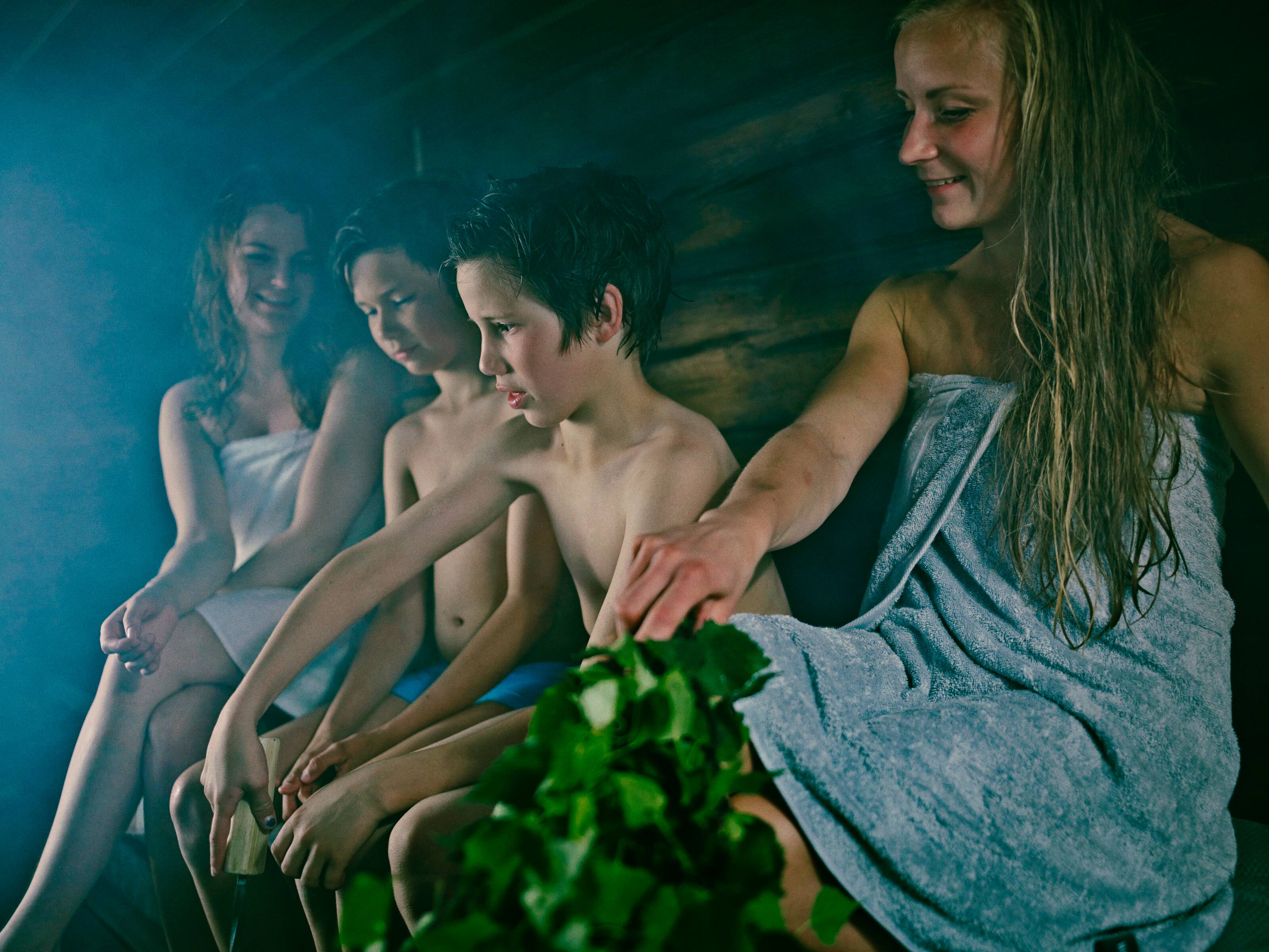 Des femmes et des enfants dans un sauna finlandais. Un enfant jette de l’eau sur un poêle de sauna
