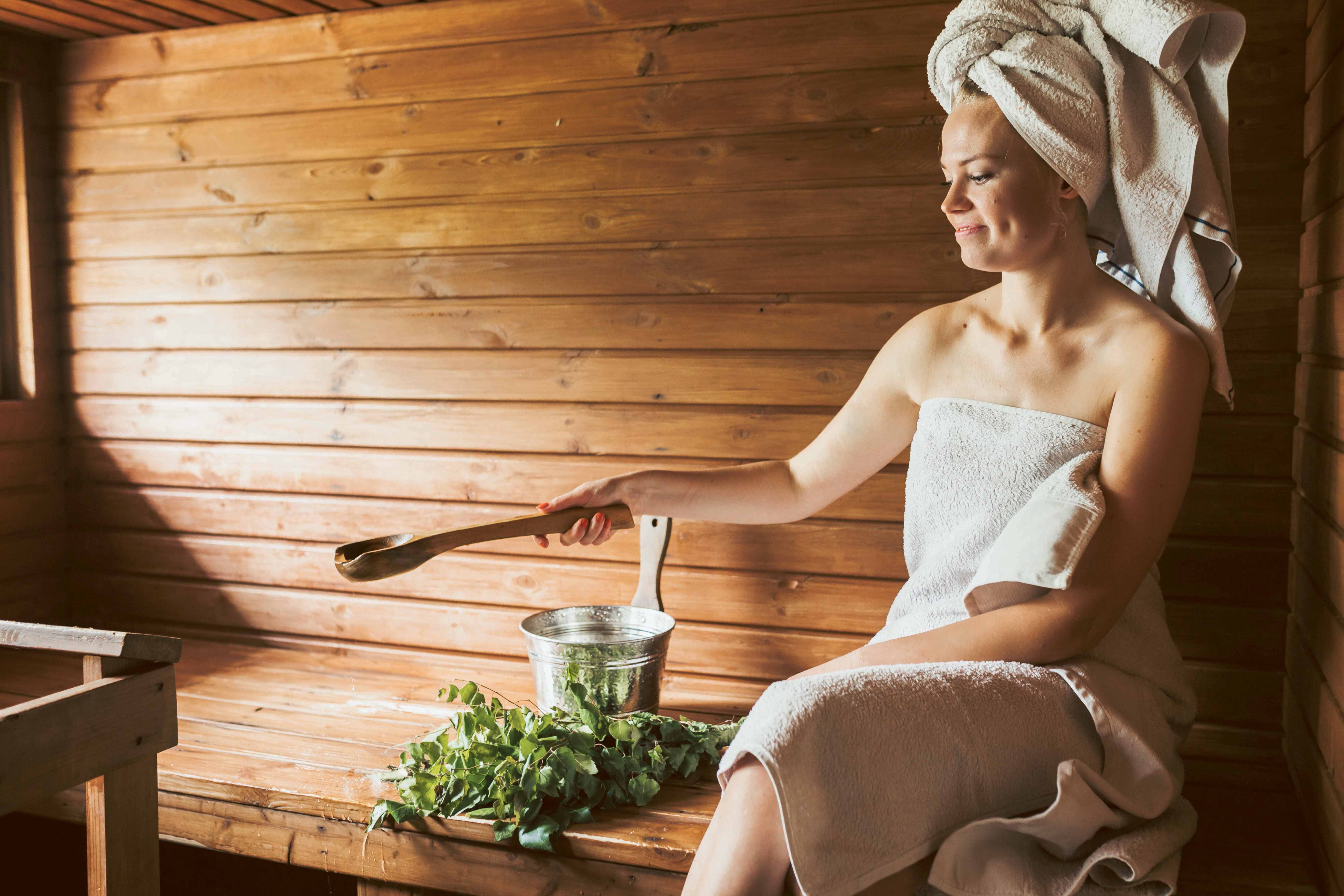 Eine mit einem Handtuch bekleidete Frau sitzt auf einer Holzbank in einer Sauna und gießt Wasser auf den Ofen